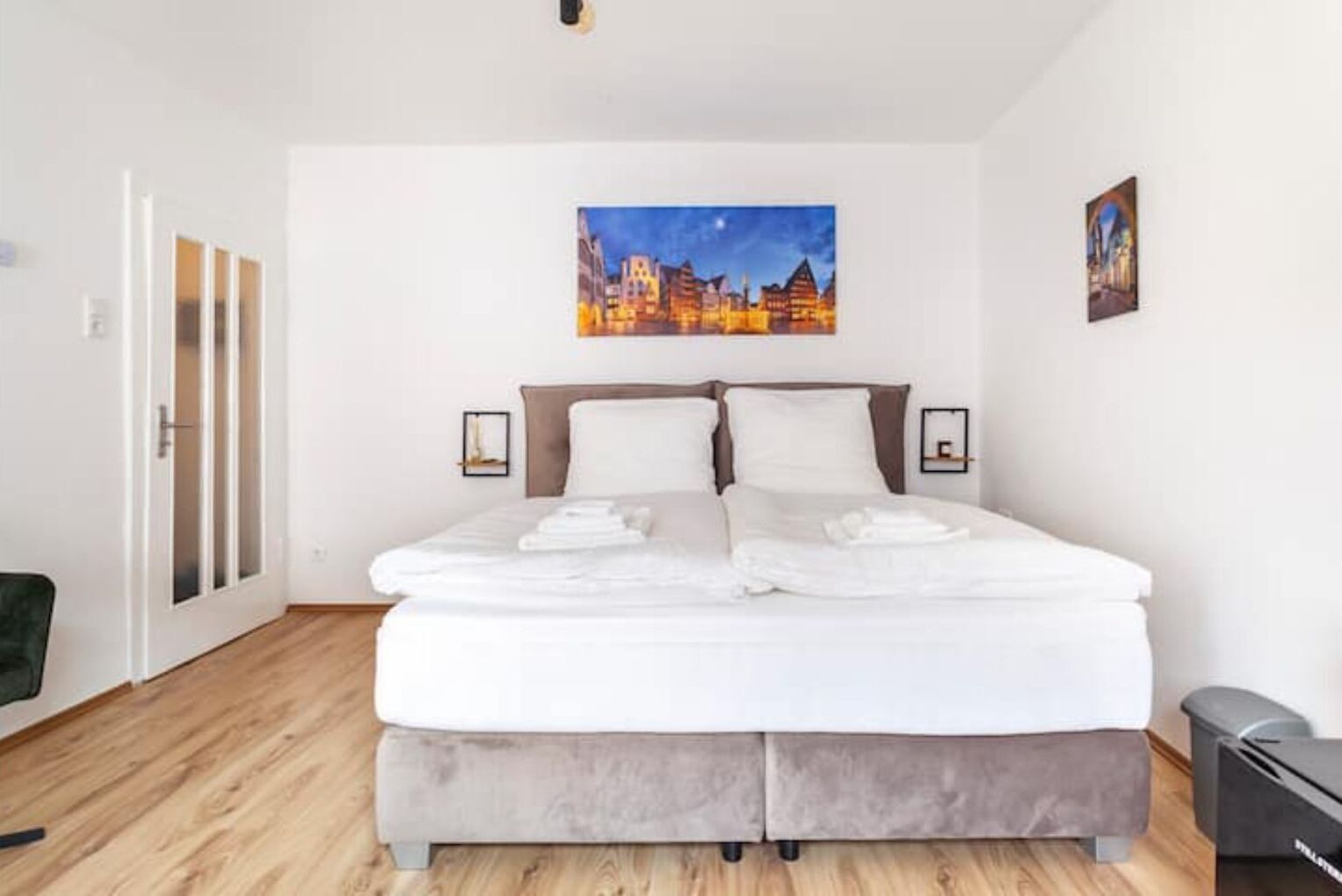 Ein modernes Hotelzimmer mit einem großen Bett, einem Stadtbild über dem Bett und Blick auf die nächtliche Stadt.