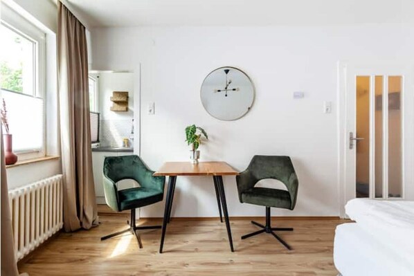 A room with a wooden floor, a round wall clock, two green chairs, a small table, and a bed. - City Apartment in Berühmter Lage (Hildesheim)