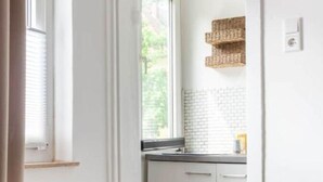 A narrow hallway leading to a kitchen with white cabinets, a window with blinds, and a radiator on the wall. - City Apartment in Berühmter Lage (Hildesheim)