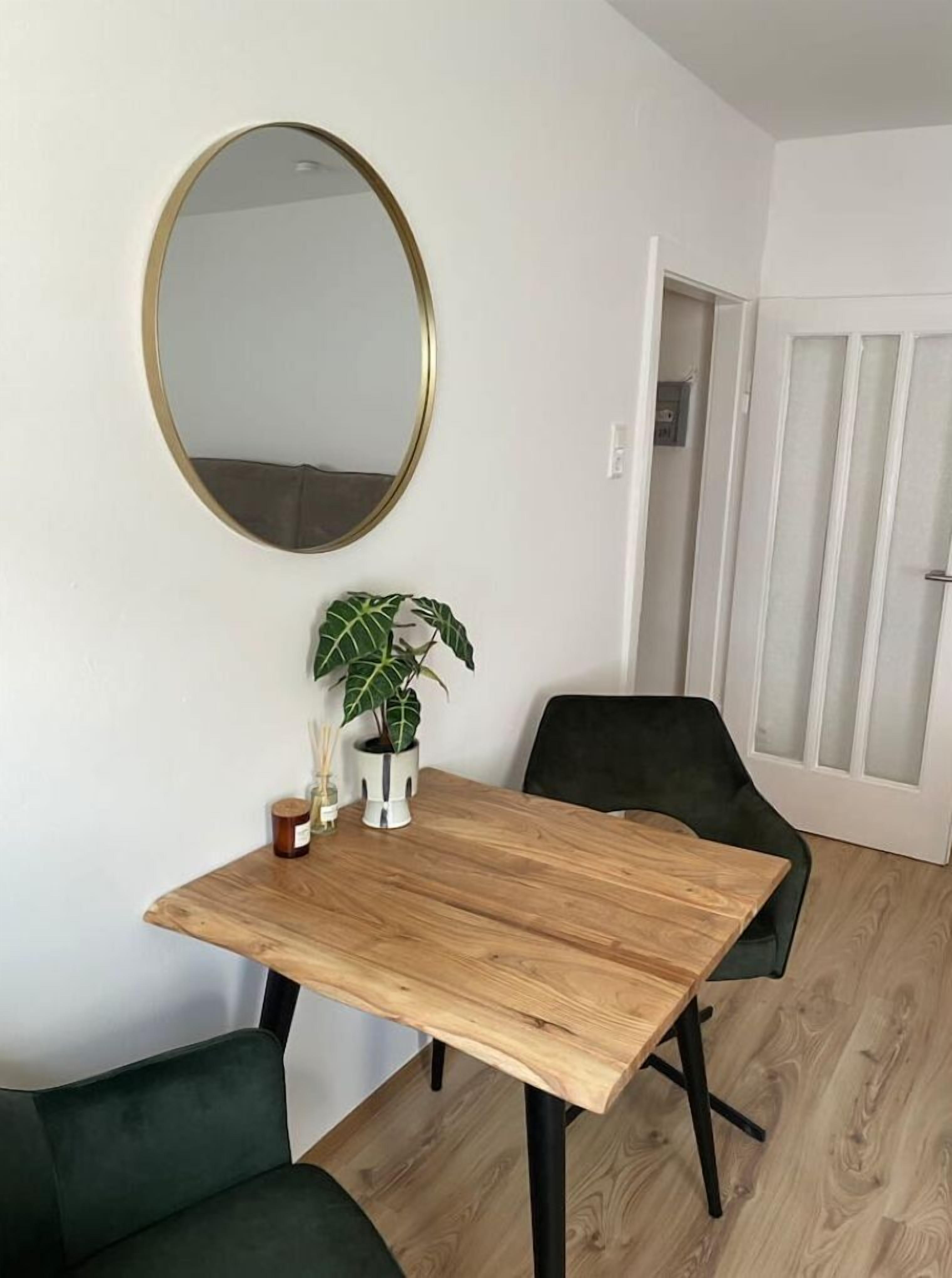 Ein Zimmer mit einem Holztisch, zwei grünen Stühlen, einem runden Spiegel und einem Blumentopf.