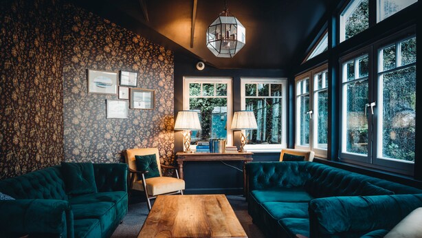 Une pièce chaleureuse avec des canapés confortables recouverts d’un tissu vert foncé, une table basse en bois et un papier peint à motifs. Des tableaux encadrés ornent le mur, une lampe est posée sur la table, et les fenêtres offrent une vue sur la verdure.