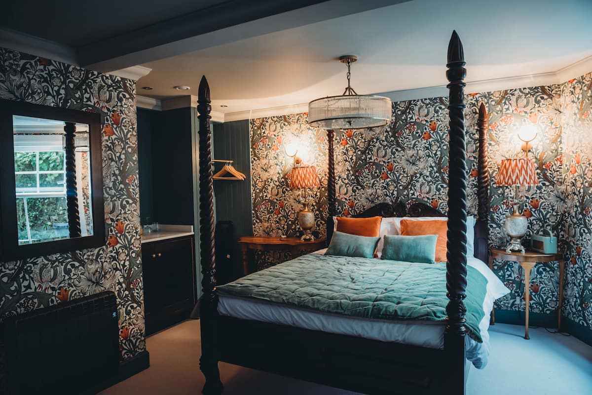 A four-poster bed with a green quilt, a chandelier, and floral wallpaper.