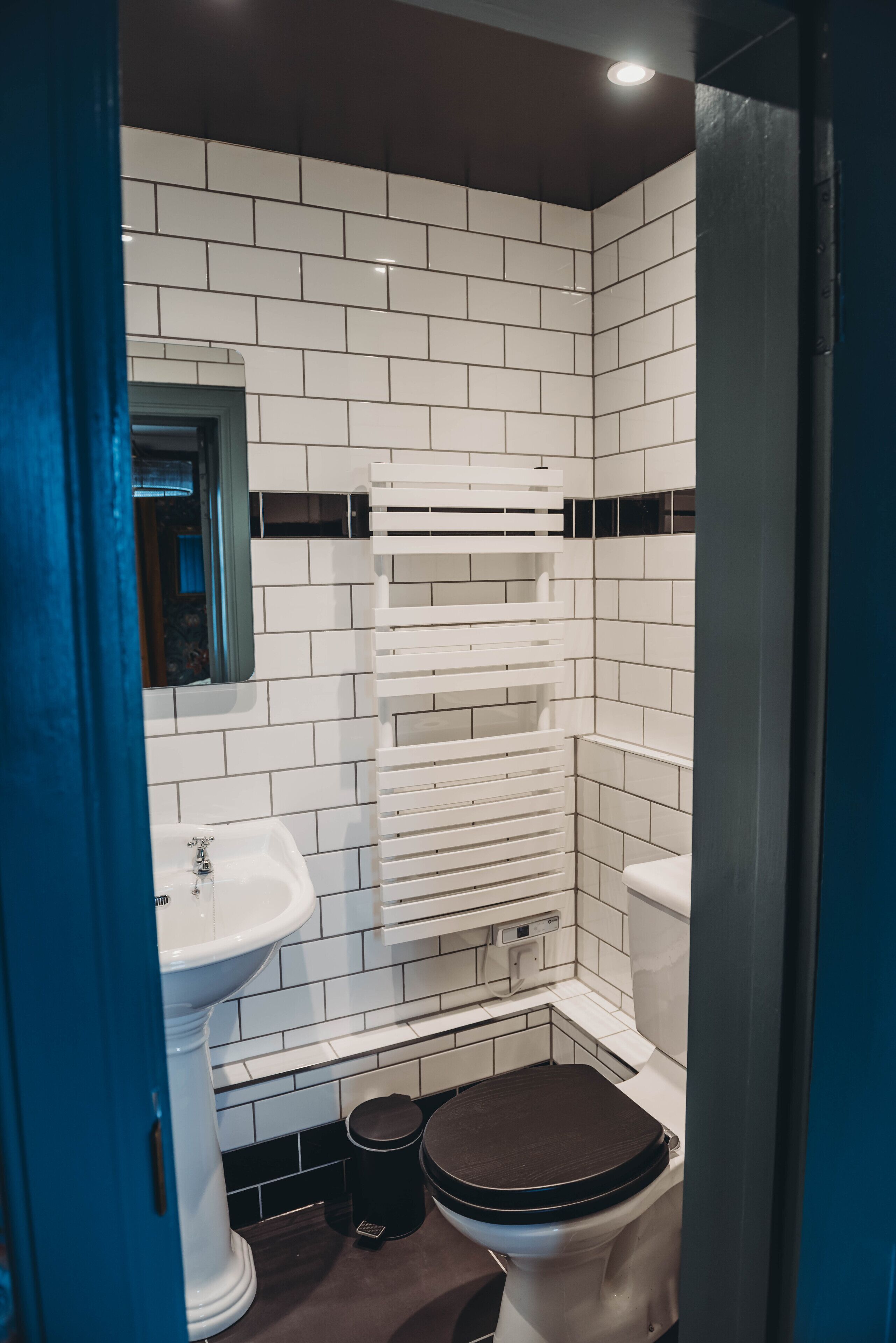 Een badkamer met witte betegelde wanden, een wit zuilwastafels, een toilet en een zwarte prullenbak.