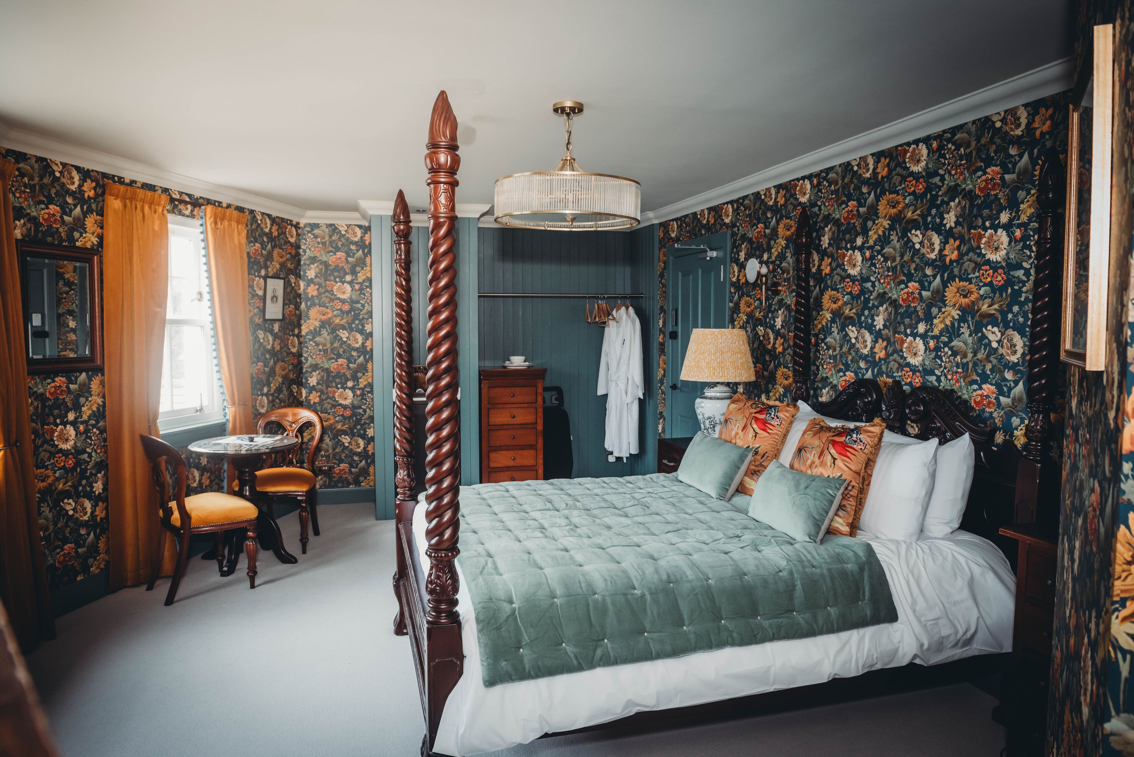 A four-poster bed with a green quilt, a wooden chair, a small round table, and a dresser with a mirror.