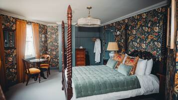 A four-poster bed with a green quilt, a wooden chair, a small round table, and a dresser with a mirror.