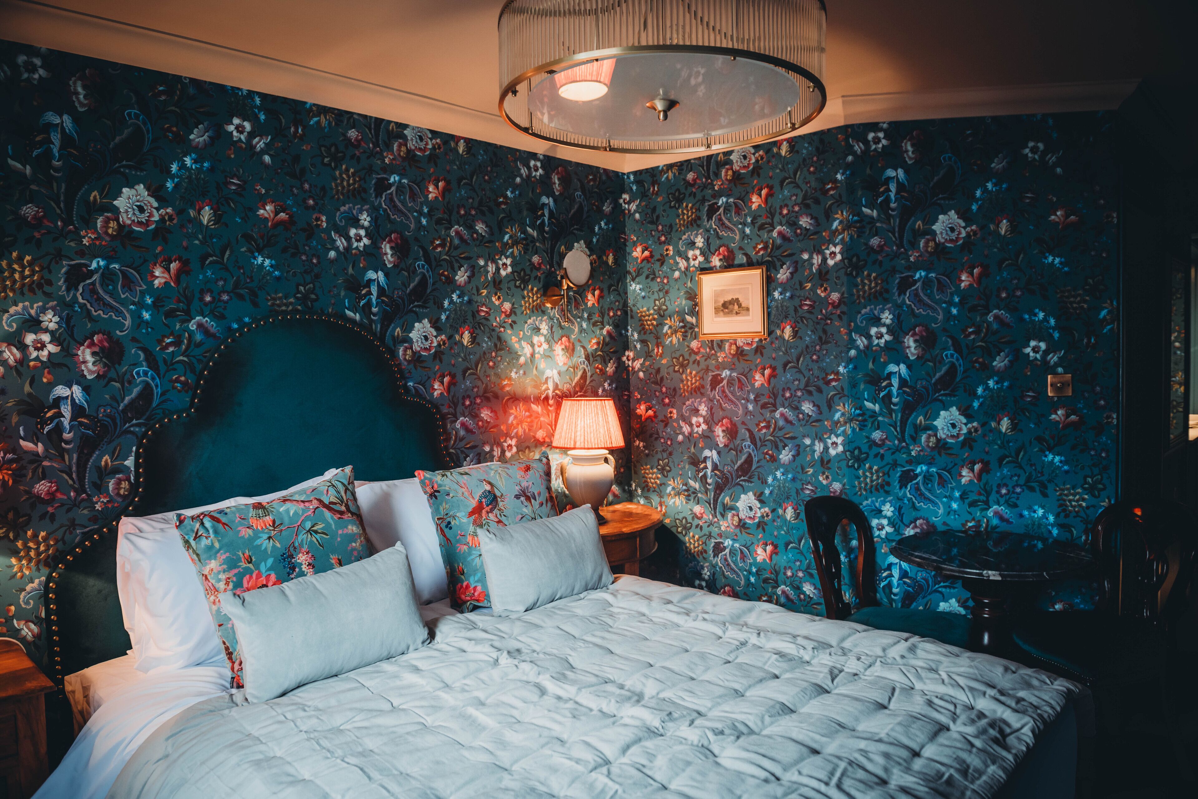 Un dormitorio con papel tapiz floral, una mesa redonda y una cama con almohadas estampadas.