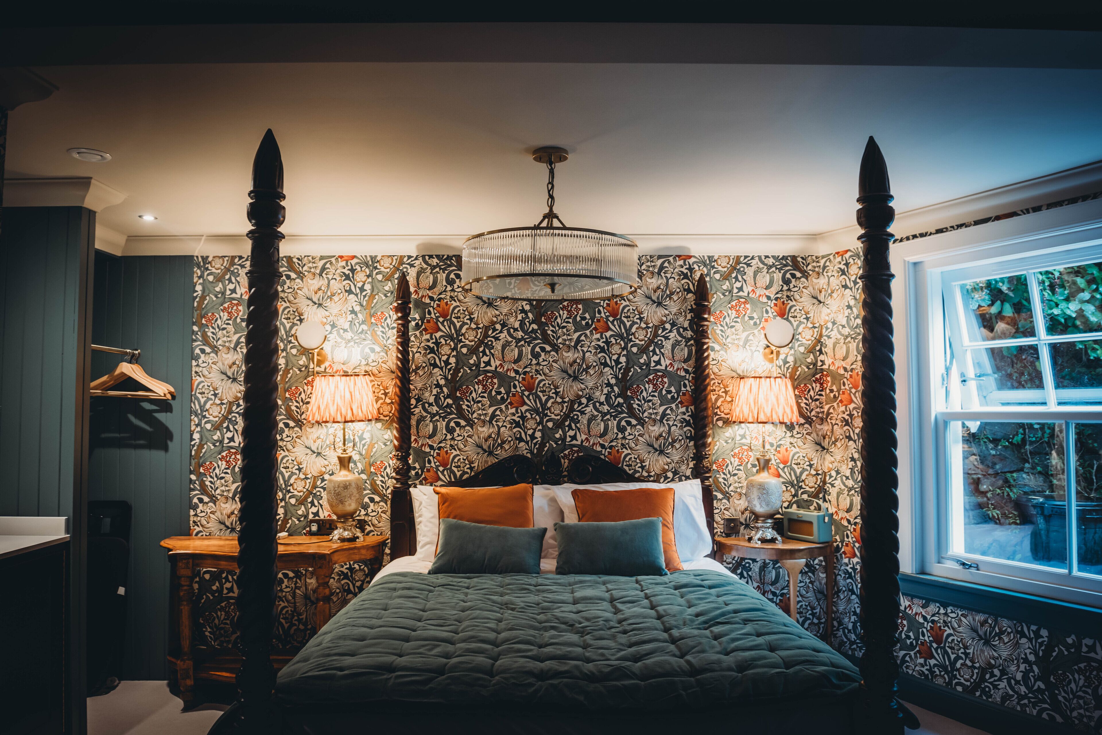 A four-poster bed with a floral wallpaper backdrop, two bedside lamps, and a chandelier.