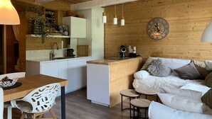 Un interior acogedor con techo de madera, una cocina con armarios blancos, un comedor con mesa y sillas, y un sofá con almohadones.