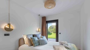 Un dormitorio con cama, almohadas, una mesita de noche, una lámpara colgante y vistas de un jardín a través de una puerta abierta.