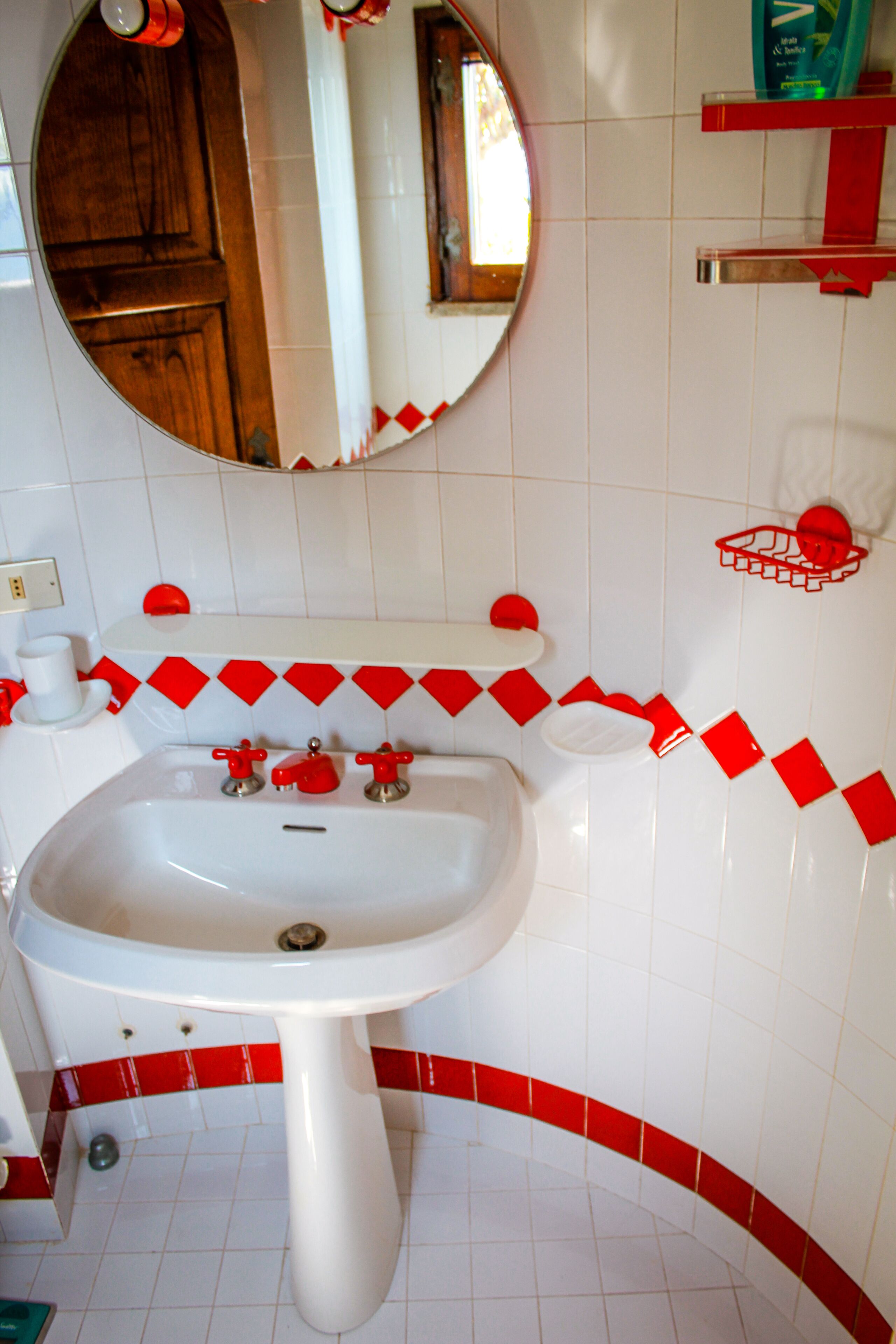 Une salle de bain avec un lavabo blanc, des murs carrelés de rouge et de blanc, une porte en bois et un miroir au-dessus du lavabo.