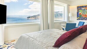 A hotel room with a bed, a desk, a chair, and a view of the sea and cliffs. - Villa Sacco - Sorrento Coast (Priora)
