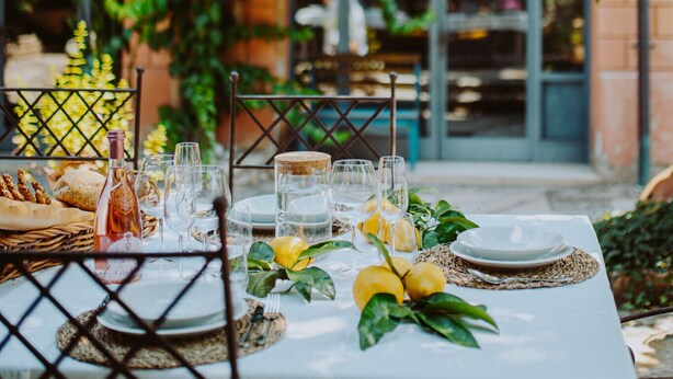 Un espace de restauration en plein air, avec une table dressée pour un repas, entouré de verdure et d’un bâtiment jaune doté de fenêtres.