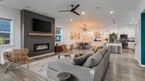 A modern living room with a fireplace, grey sofa, wicker chairs, and a dining area with a wooden table and chairs.