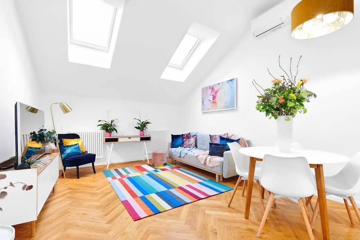 Un appartement moderne et bien éclairé, doté d’un plafond de style industriel, d’un coin repas avec une table et des chaises blanches, d’un tapis rayé aux couleurs vives et d’un coin salon confortable avec un canapé et des coussins.