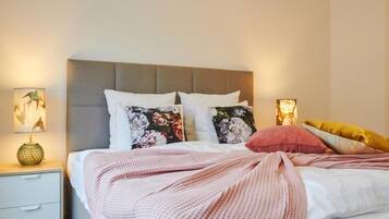 Una cama bien hecha con cabecera gris, almohadas blancas y florales, y una manta rosa. Hay dos lámparas de noche con diseños florales.