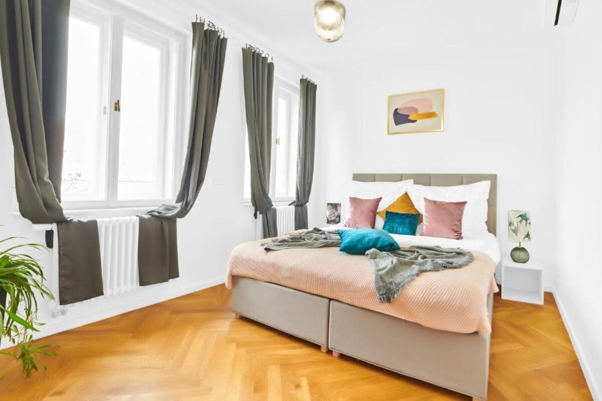 Un dormitorio con cama, cortinas, un radiador, una planta y un cuadro en la pared.