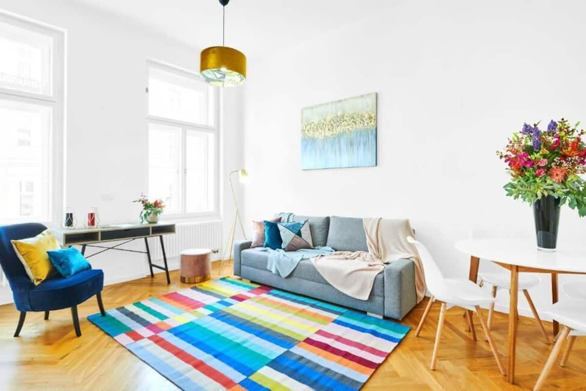Un salon moderne avec un canapé gris, un fauteuil bleu, un tapis rayé aux couleurs vives, une table à manger blanche et un arrangement floral éclatant.