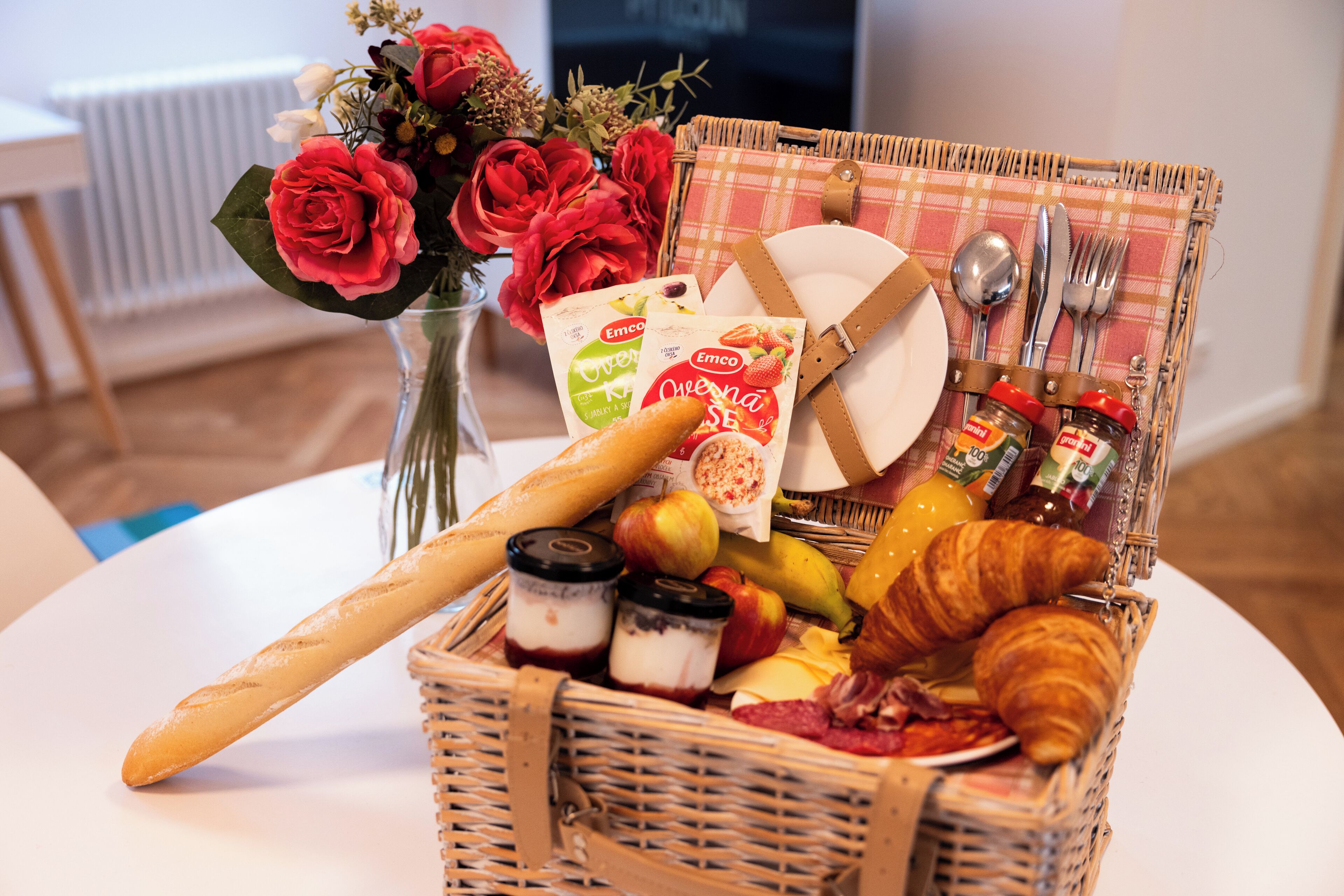 A wicker basket with food items, a baguette, a croissant, and a vase of red flowers.