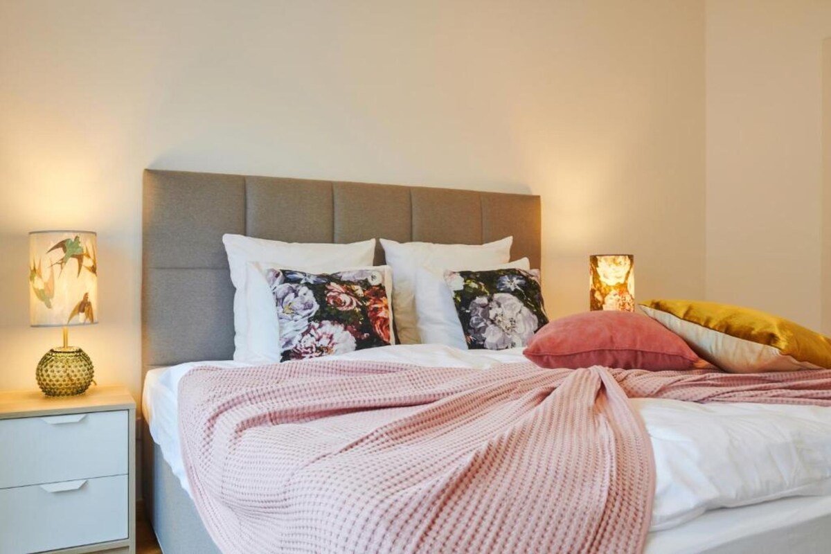 Una cama bien hecha con cabecera gris, almohadas blancas y una colcha rosa. Hay dos lámparas de noche con diseños florales.