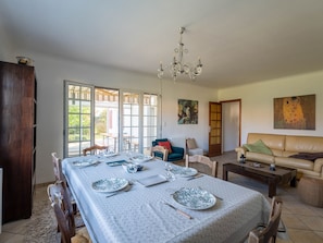 Un comedor con una mesa dispuesta para cuatro, una araña y un sofá en una habitación luminosa con ventanas amplias.