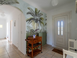 Un couloir orné d’une fresque représentant des feuilles tropicales, un meuble à vin en bois, une porte blanche avec des vitraux et une chaise blanche.