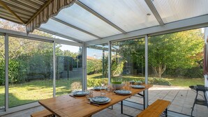 A conservatory with a wooden table set for dining, large glass doors, and a view of a garden. - Cozy Family Home in Parentis-en-Born: 3BR, WiFi, Large Garden, Near Beach & Lake (Parentis-en-Born)