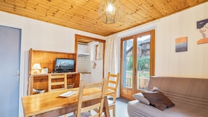 A cozy room with a wooden ceiling, a dining table with chairs, a sofa, and a TV on a wooden cabinet. - Cozy 1BR Apartment, Balcony, Pool, Close to Serre Chevalier Slopes, Free Wi-Fi & Parking (Saint-Chaffrey)