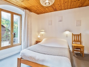 Una habitación con techo de madera, una cama con colchón blanco, una silla de madera, una mesita de noche con lámpara y una ventana con cortinas.