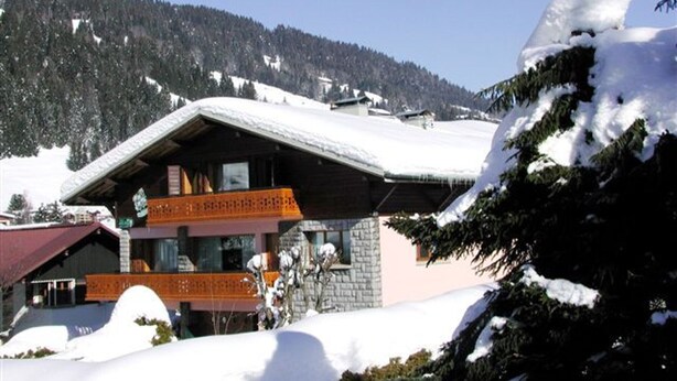 Een chaletachtige woning met een schuin dak, houten balkon en gevel van natuursteen, omgeven door besneeuwde bergen.