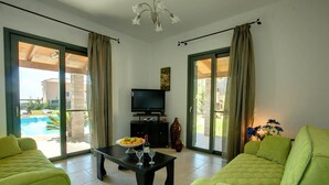 Ein Wohnzimmer mit Blick auf einen Pool, mit grünen Sofas, einem Fernseher und einem Esstisch mit Obst und Wein.