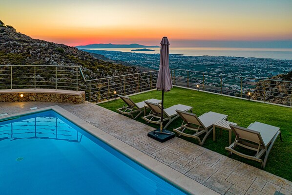 A pool with lounge chairs and an umbrella, overlooking a coastal cityscape at sunset.