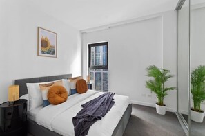 Ein modernes Schlafzimmer mit Bett, Kissen, einer Nachttischlampe, einem Fenster mit Stadtblick und zwei Blumentöpfen.