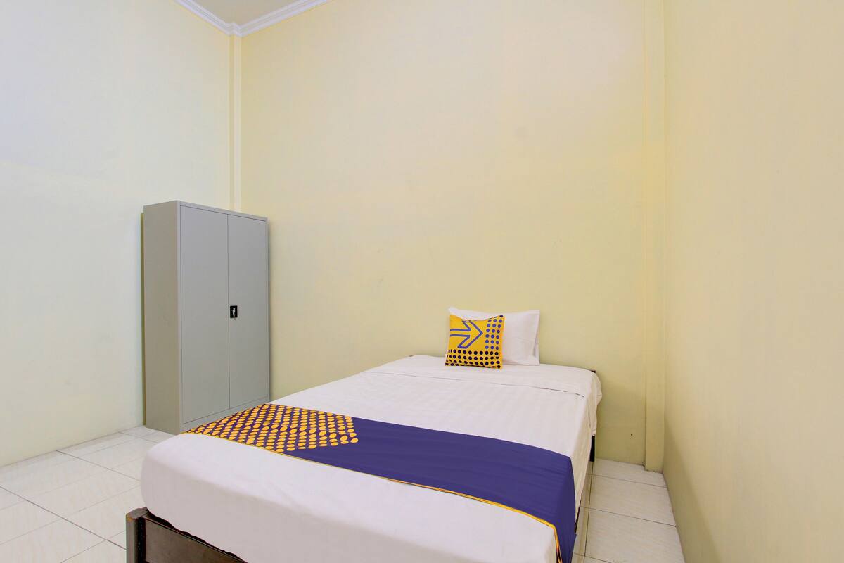A small, neatly made bed with a white and blue bedspread, a single pillow, and a wardrobe against a plain wall.