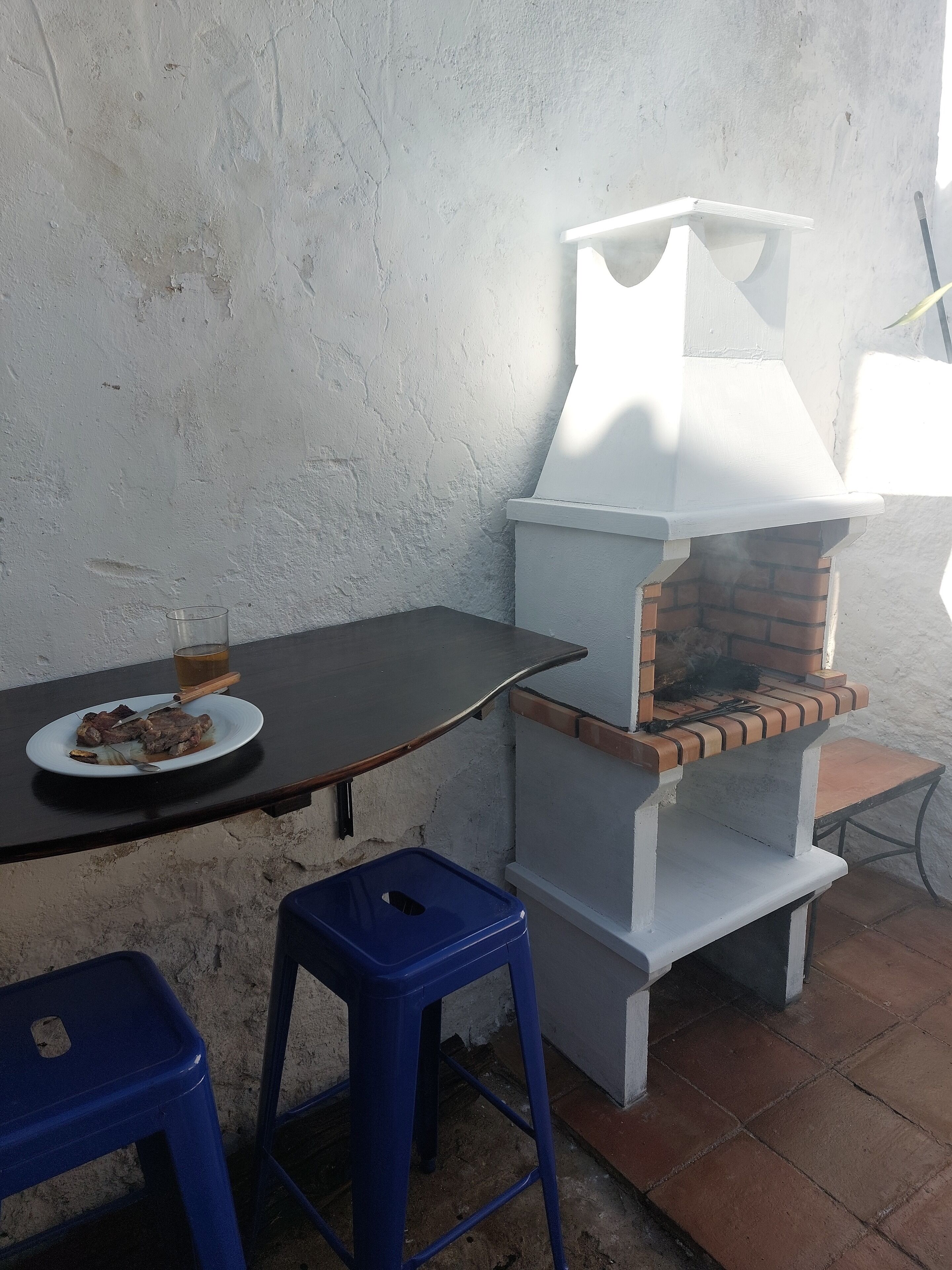 Un espacio de comedor al aire libre rústico, con un horno de ladrillo, una mesa y taburetes azules.
