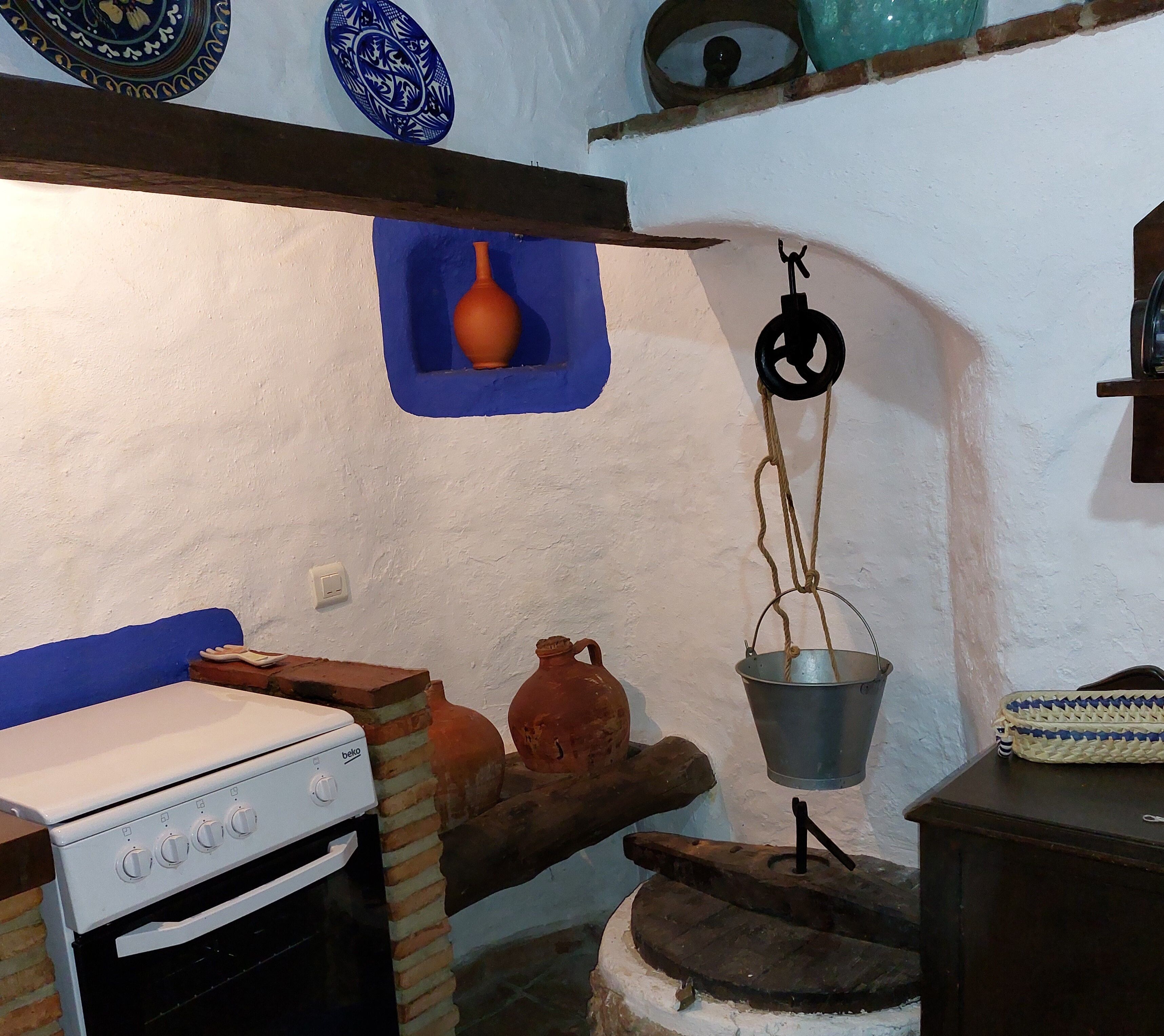 A rustic kitchen with a white stove, clay pots, and a hanging metal bucket.