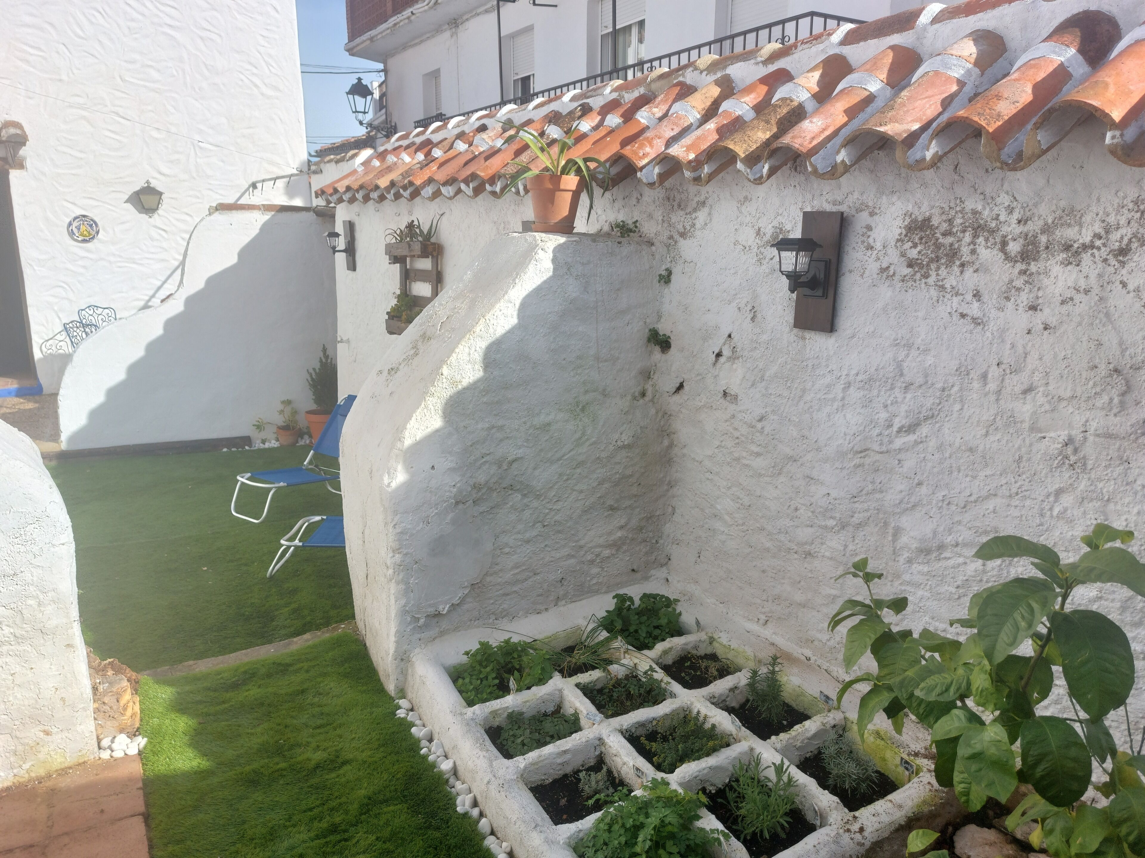 Un bâtiment blanc avec un toit recouvert de tuiles, un petit jardin avec de l’herbe et des plantes, et une chaise bleue.