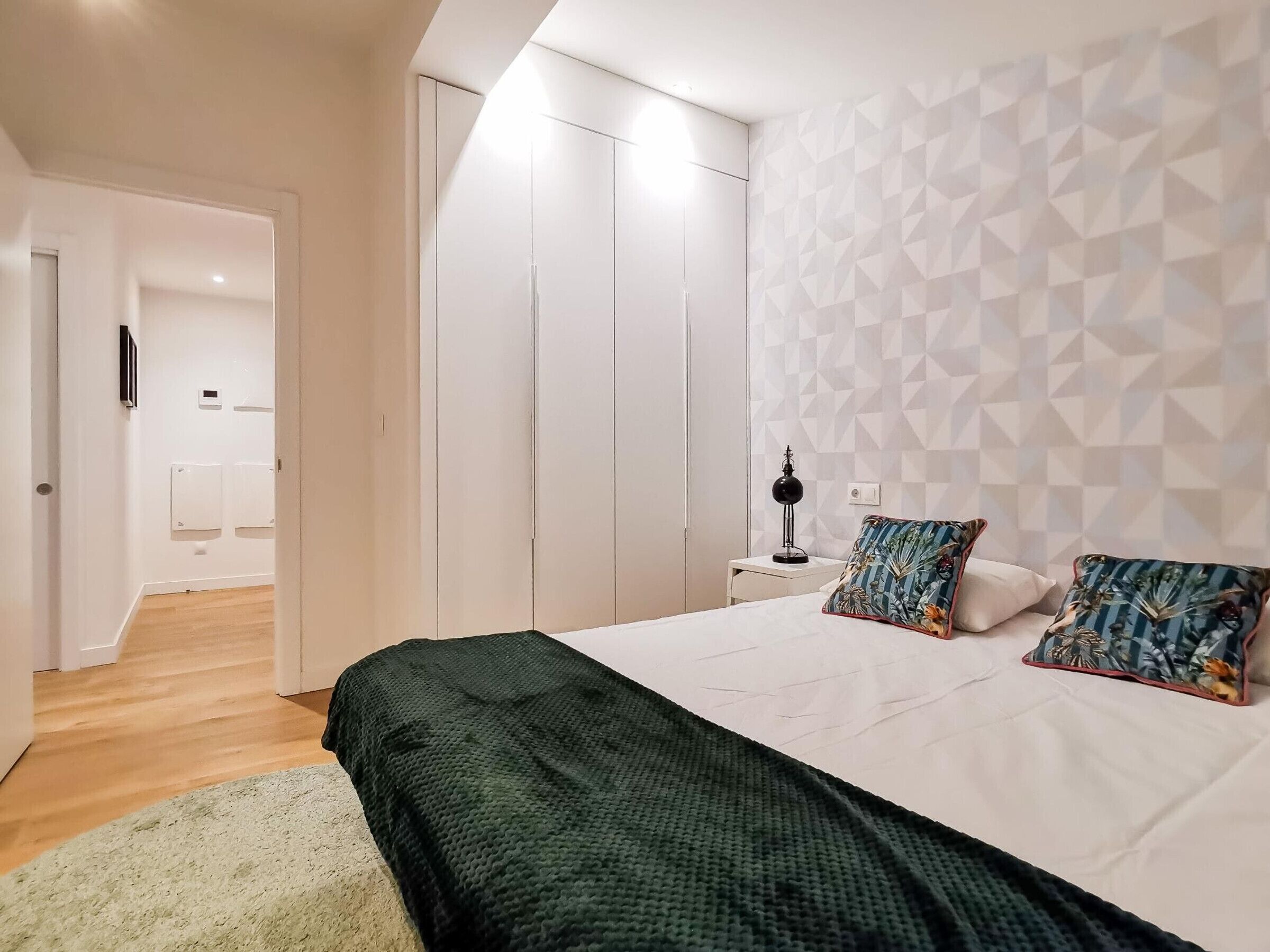 Un dormitorio con cama, almohadas, una mesita de noche, una lámpara y un papel tapiz con diseño geométrico.