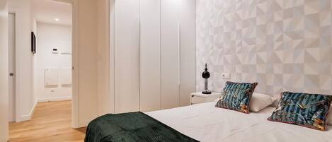Un dormitorio con cama, almohadas, una mesita de noche, una lámpara y un papel tapiz con diseño geométrico.