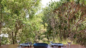 A table set for dining with plates, bowls, and wine glasses, surrounded by dense foliage. - Twin Waters Seaside Resort Life (Twin Waters)
