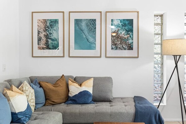 A living room with a grey sofa, multiple cushions, and three framed abstract paintings on the wall.