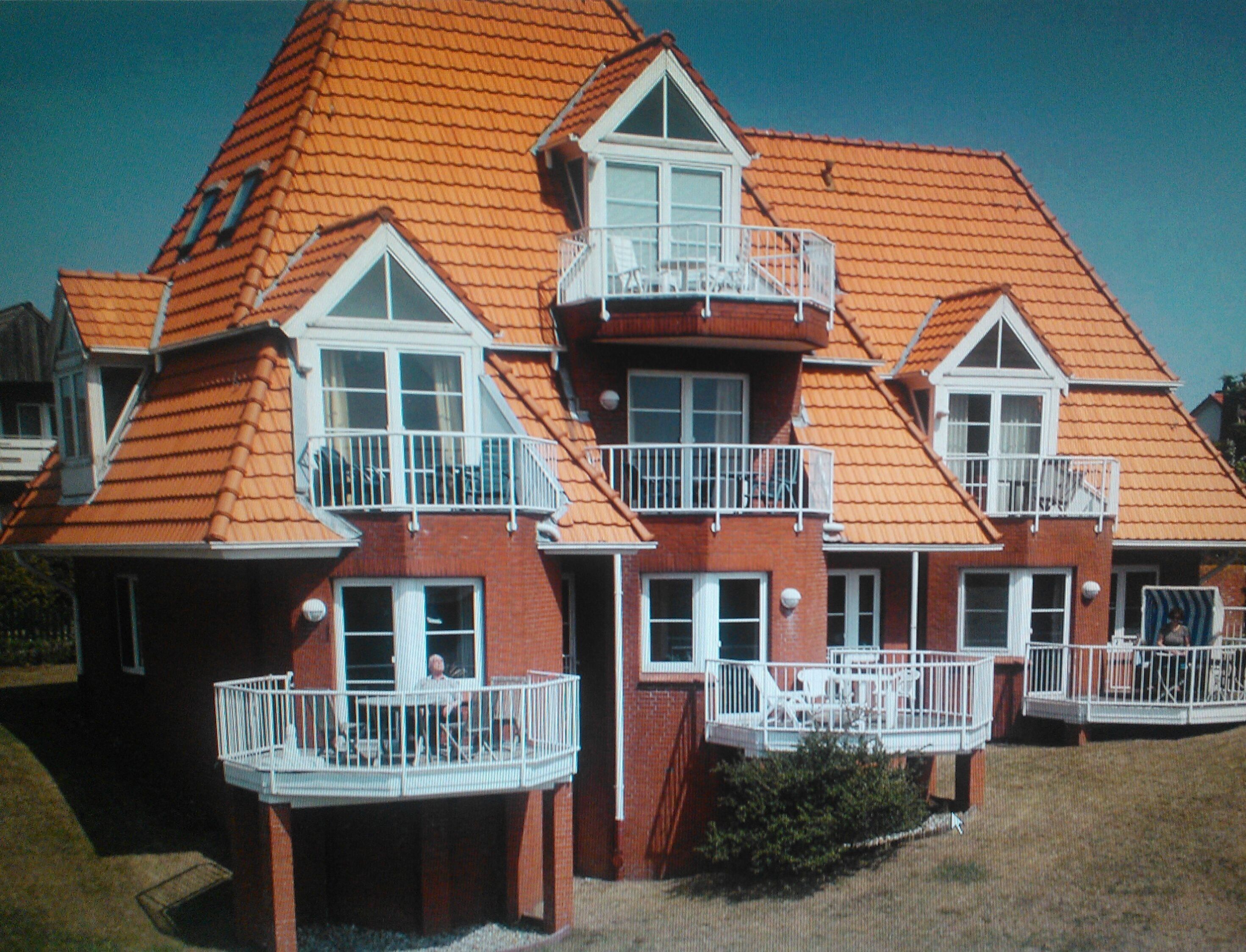 Una casa de varios pisos con fachada de ladrillo rojo y tejas del color naranja. Tiene varios balcones con barandillas blancas y un pequeño porche delantero con mobiliario exterior.