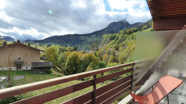 A balcony with a view of a mountain landscape, a wooden building, and a metal chair.