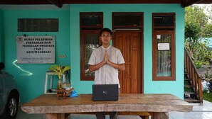 A person standing at a wooden table with a laptop, in front of a building with a signboard. - OYO 93248 Villa Syariah Astuti Lestari (Parongpong)