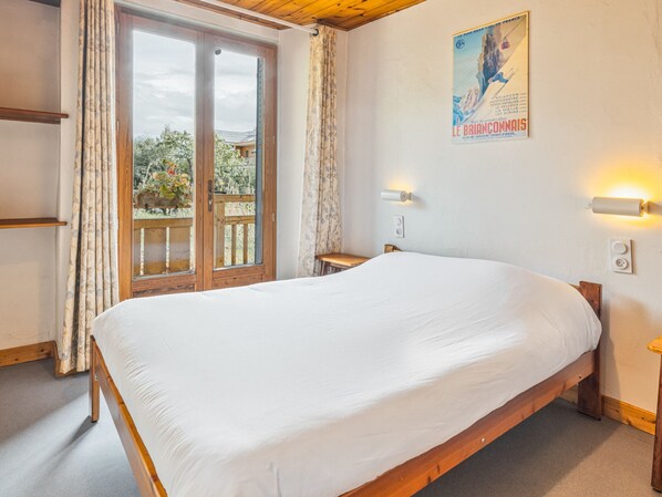A bedroom with a wooden bed, a window with curtains, a poster on the wall, and a small table. - Cozy 1BR+Alcove in Serre Chevalier with Pool, WiFi, Private Parking, Near Slopes (Saint-Chaffrey)