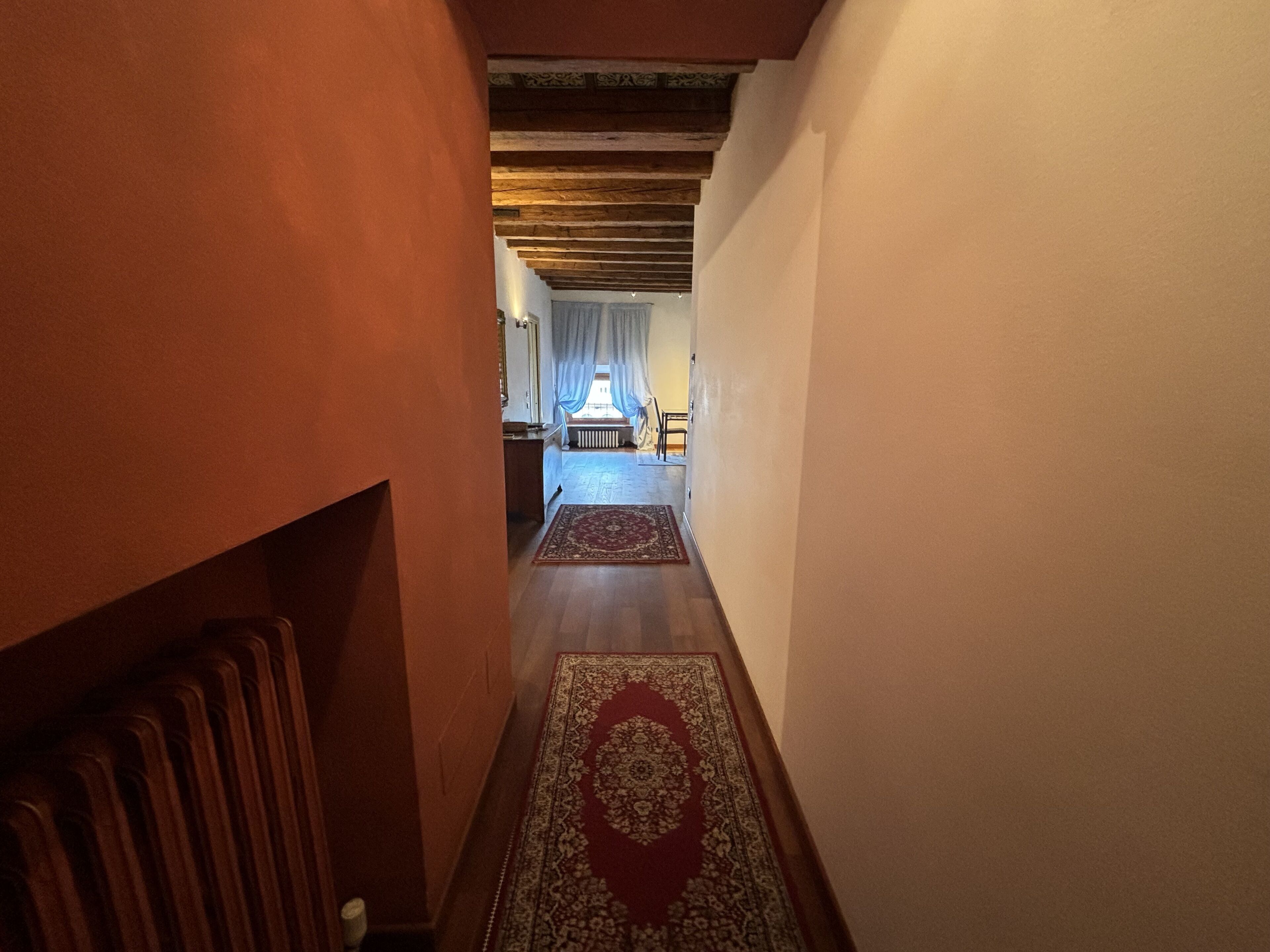 A narrow hallway with wooden floors and a patterned rug leading to a room with a bed and a window with curtains.