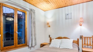Un dormitori amb sostre de fusta, una finestra gran amb cortines, un llit amb almoades blanques i una cadira de fusta.