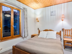 A bedroom with a wooden ceiling, a large window with curtains, a bed with white pillows, and a wooden chair. - Charming 1-BR Apartment for 4 with Pool, Wi-Fi & Parking, Near Ski Slopes (Saint-Chaffrey)