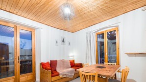 Una habitació acollidora amb sostre de fusta, un sofà de fusta amb coixins, una taula de dinar amb cadires i una finestra gran amb cortines.