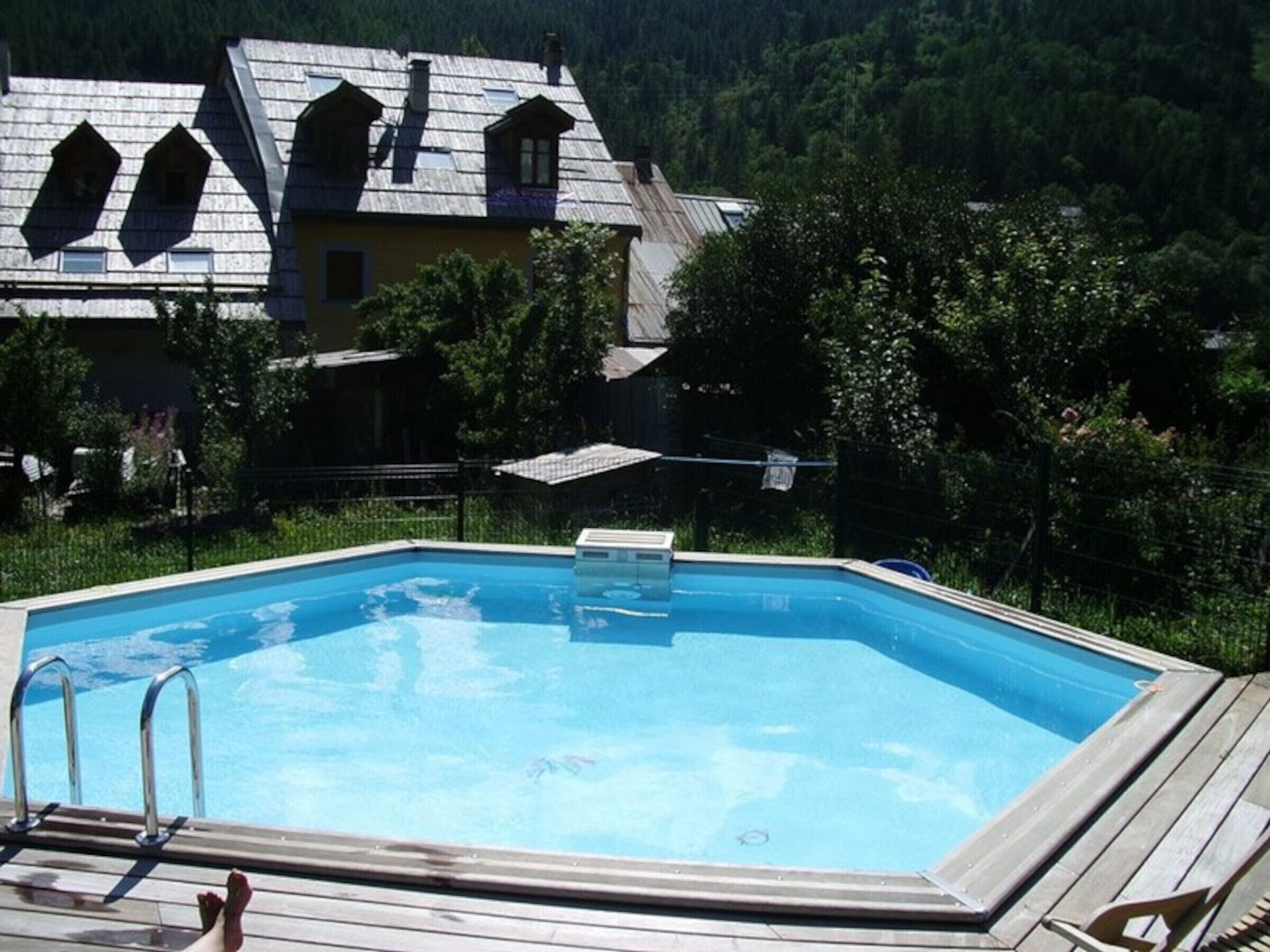 A swimming pool with a wooden deck and metal ladder, surrounded by lush greenery and a house with a sloped roof.