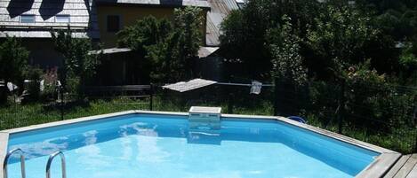 A swimming pool with a wooden deck and metal ladder, surrounded by lush greenery and a house with a sloped roof.